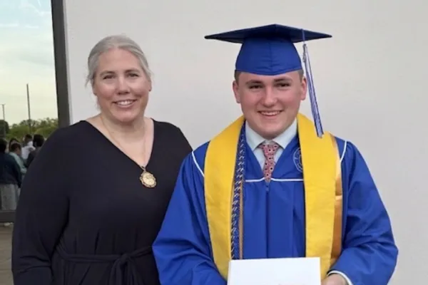 Photo of high school graduate in blue cap and gown with yellow stole, holding diploma and standing with his mother who is in black. 