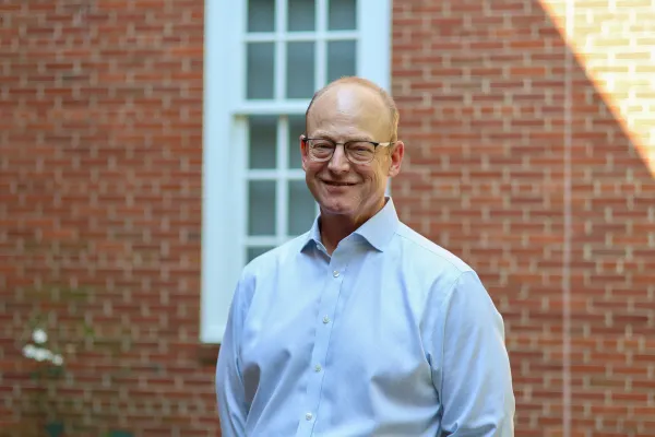 Photo of man in light blue button up shirt against a brick building background 