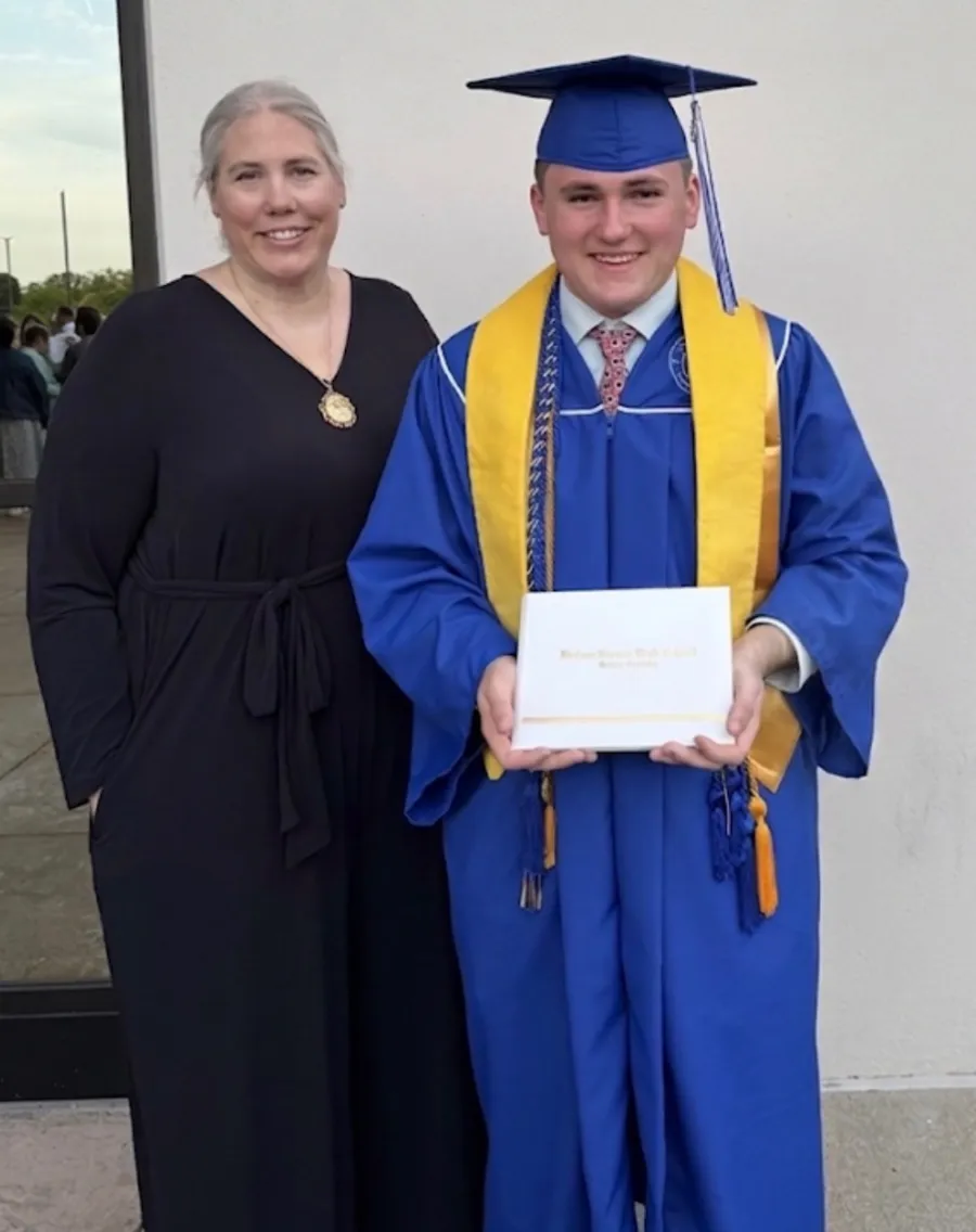 Photo of high school graduate in blue cap and gown with yellow stole, holding diploma and standing with his mother who is in black. 